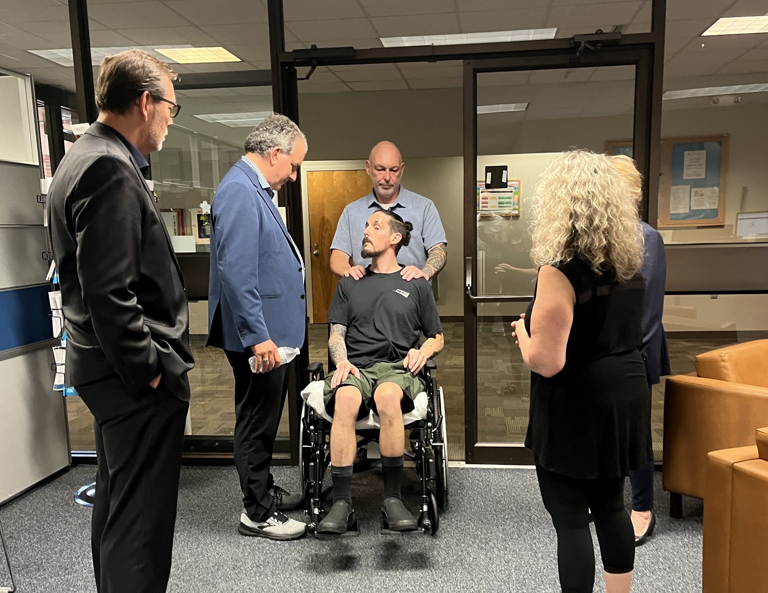 U.S. Representative Jimmy Patronis, wearing a blue blazer, and VetCV Foundation Board Member Niels Andersen in a dark suit stand in an office setting, looking respectfully toward a disabled veteran seated in a wheelchair. The veteran’s father stands behind the veteran with his hands placed supportively on his shoulders, while the veteran’s mother stands to the right observing the interaction.
